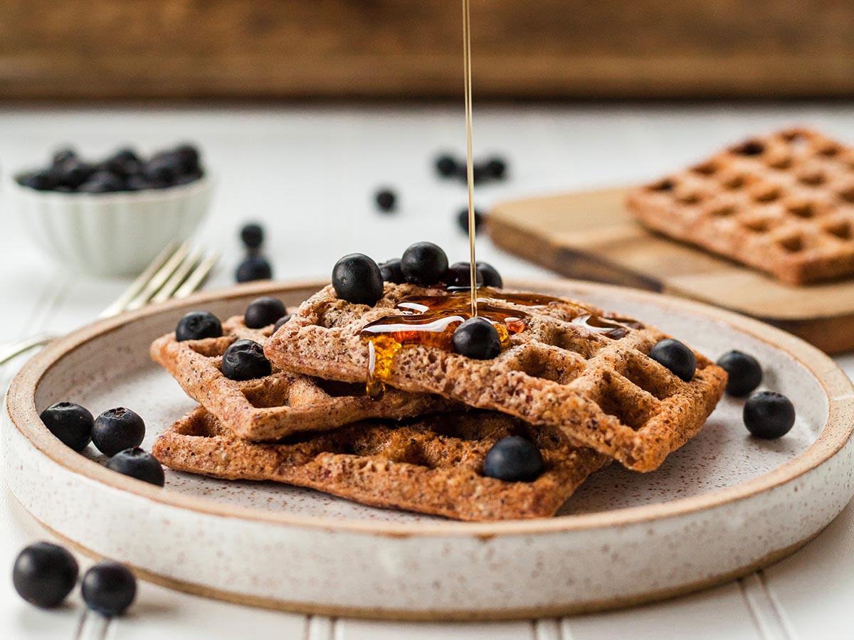 Maple syrup being poured onto whole grain waffles with fresh blueberries